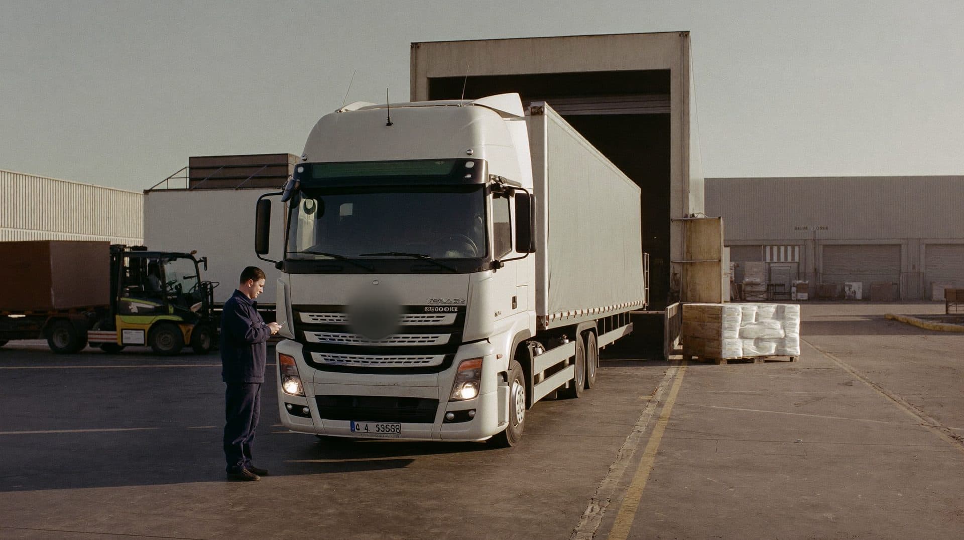 Sabah ışığında yükleme rampasında dorselerin yanında telefonuna bakan sürücü ve forklift operatörü